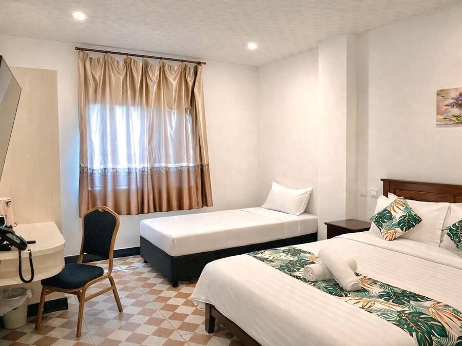 A hotel room with a checkered floor, a desk with a telephone, a chair, two beds with different bedding, and a window with curtains.