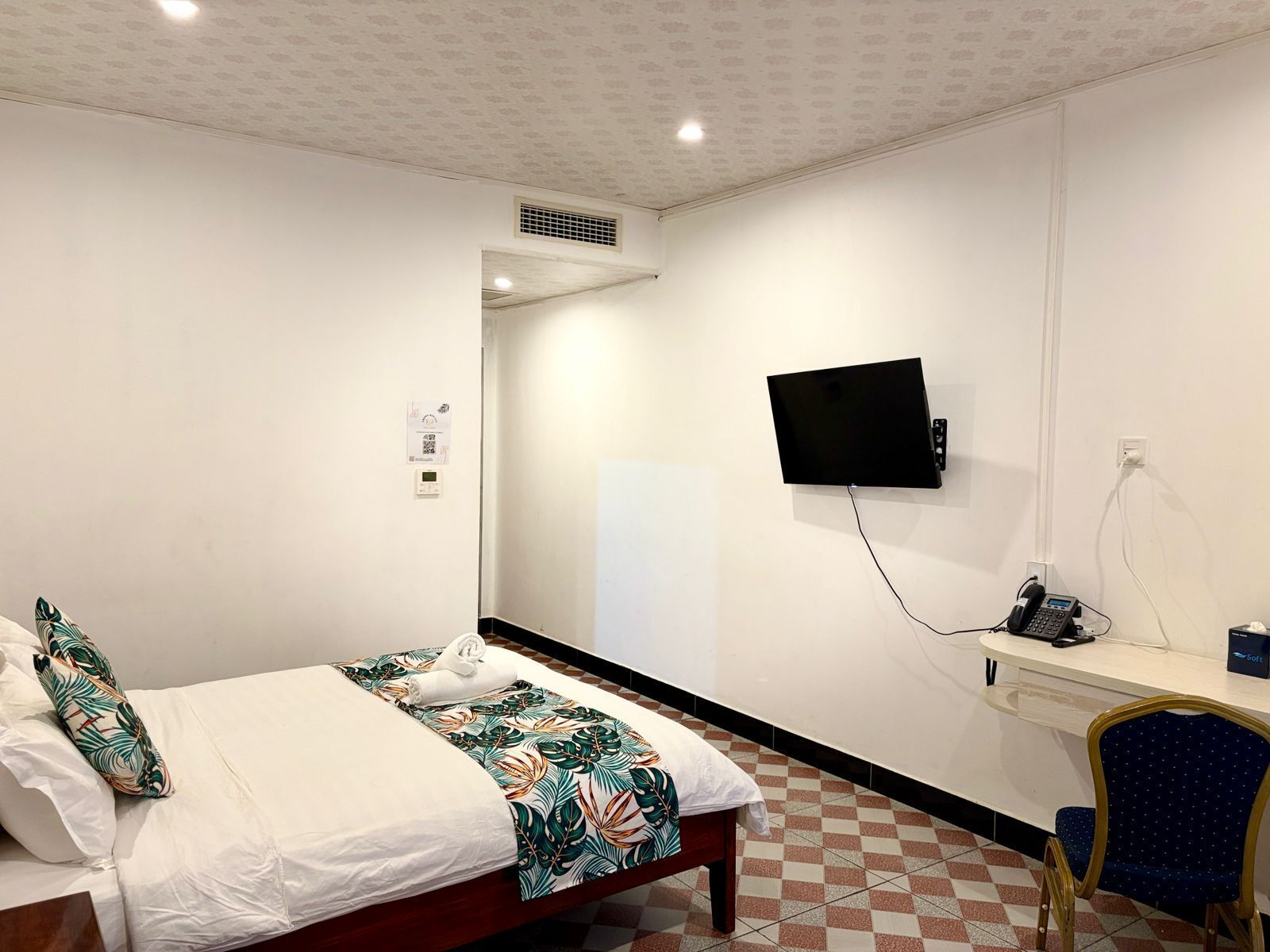 A hotel room with a bed, a desk with a telephone, a flat-screen TV mounted on the wall, and a checkered floor.