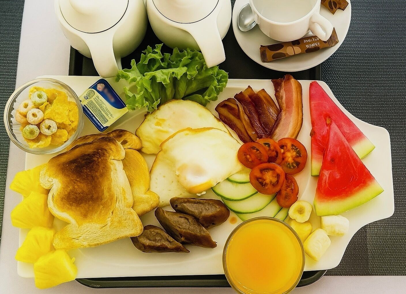Una bandeja de desayuno con tostadas, huevos, tocino, tomates, rebanadas de pepino, sandía y un vaso de jugo de naranja.