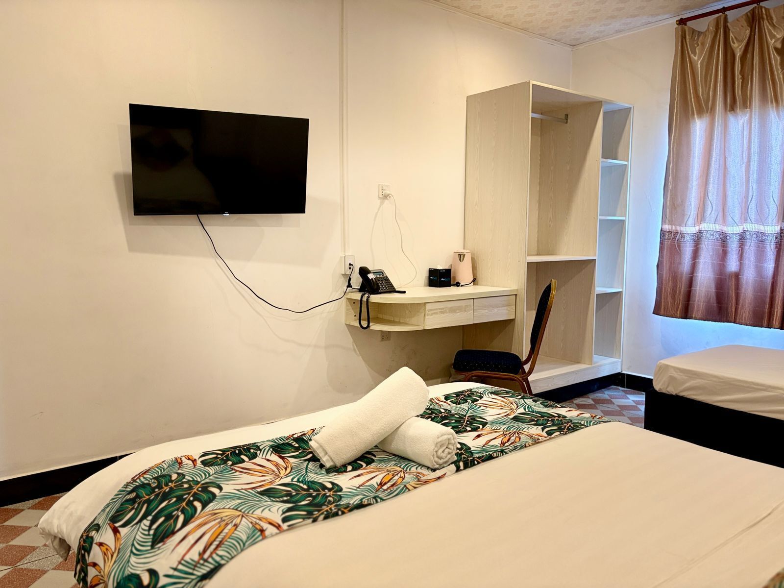 A hotel room with a bed, a TV mounted on the wall, a desk with a phone and a lamp, a chair, and a wardrobe.