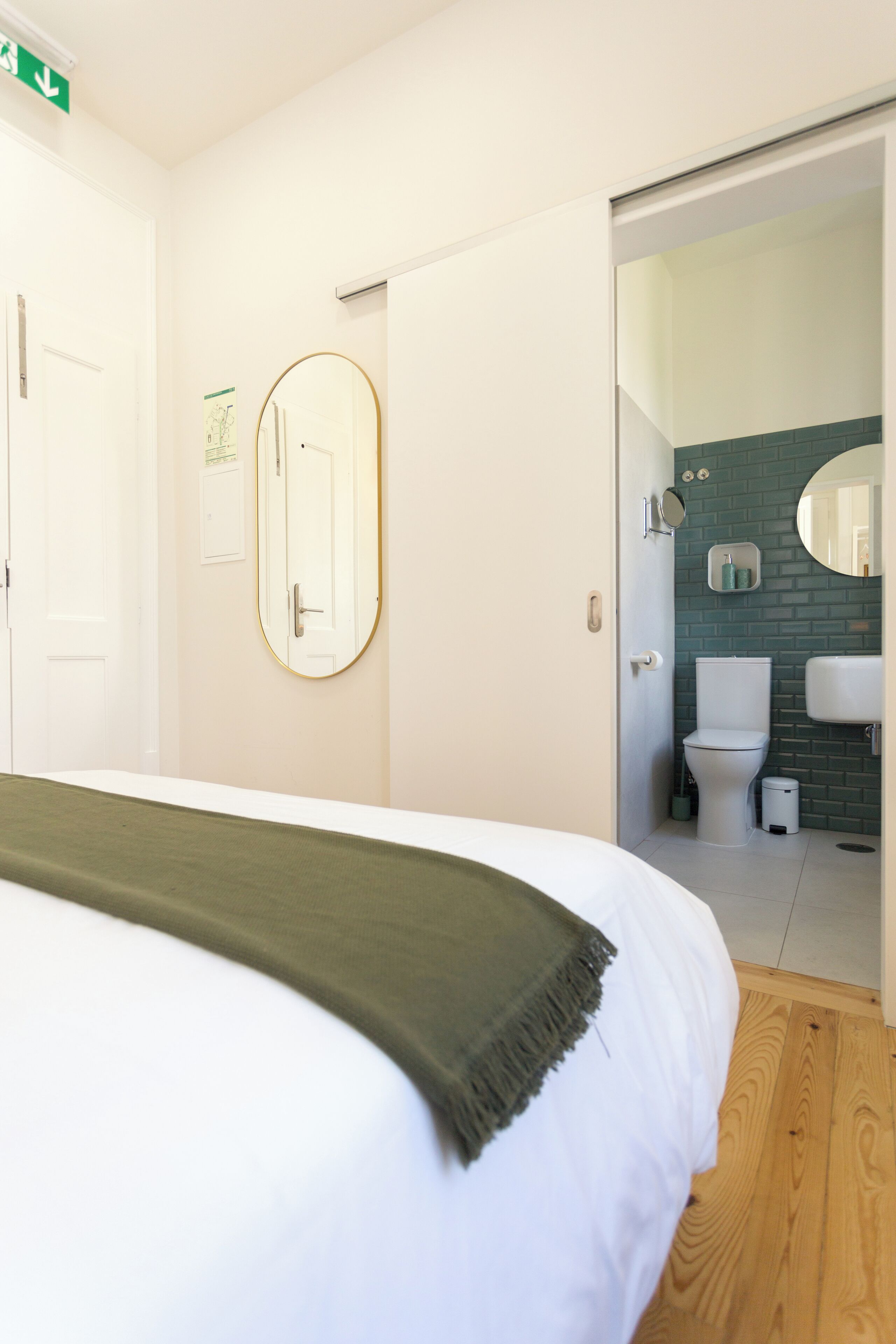 Ett hotellrum med en säng, en spegel, en dörr och utsikt in i badrummet.