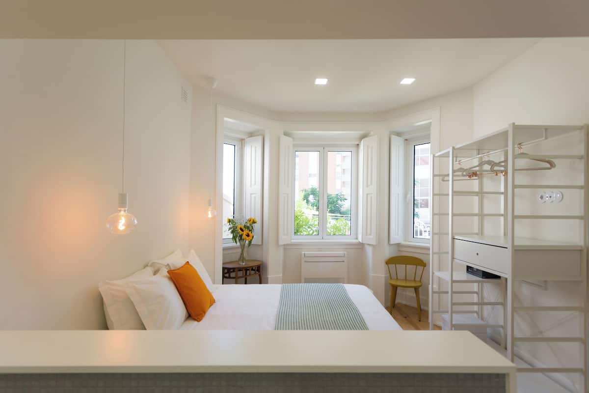 A bedroom with a bed, a desk, and a chair. There is a window with a view of buildings outside.
