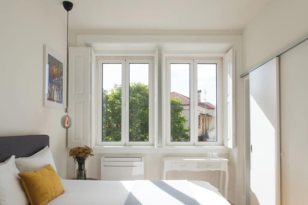 A bedroom with a large bed, two windows, a bedside table with a vase, and a view of greenery outside.