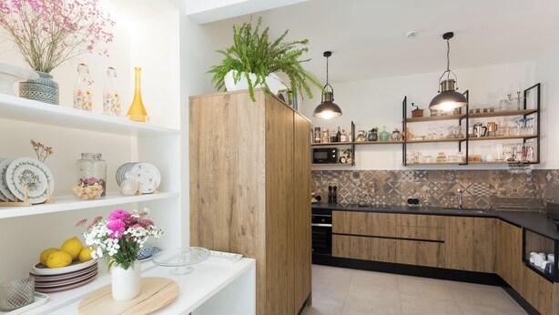 Une cuisine moderne avec des armoires en bois, un comptoir noir et un pare-feu en carreaux. On y trouve des étagères ornées d’objets décoratifs et un pot de fleurs posé sur le comptoir.