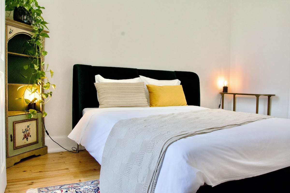 A neatly made bed with white and yellow pillows, a dark headboard, and a bedside table with a lamp.