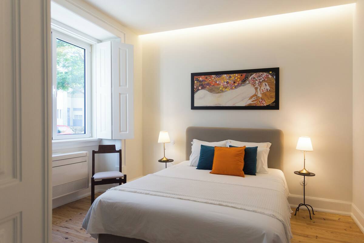 A bedroom with a large bed, two bedside lamps, a chair, a radiator, and a window with a view of the outside.