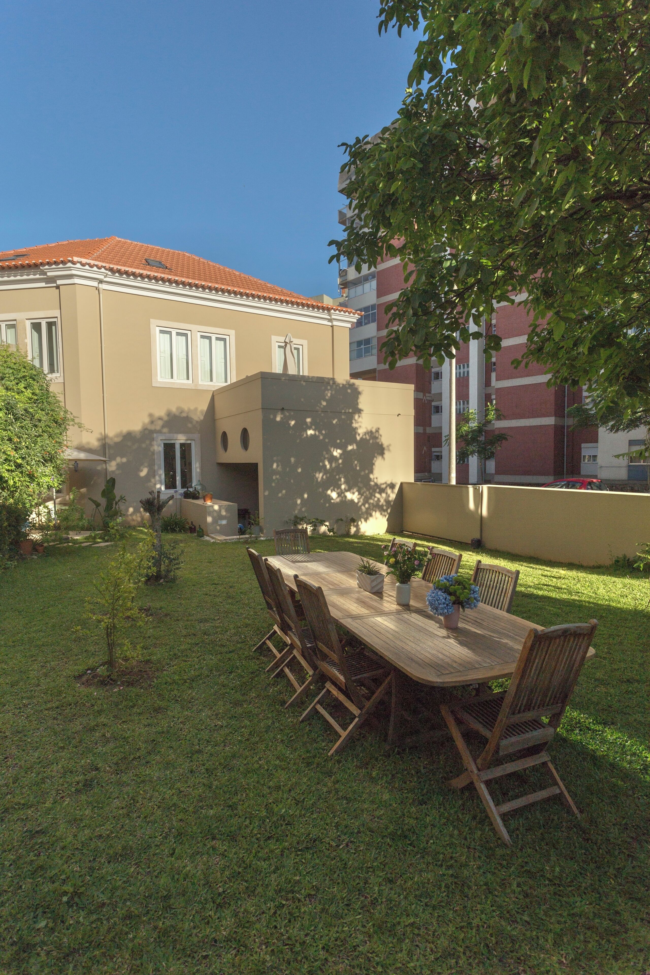 A patio with a wooden table and chairs, surrounded by a well-maintained lawn and a building with a red-tiled roof.