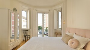Une chambre avec un grand lit, une armoire et une chaise. On peut voir des fenêtres et un balcon.