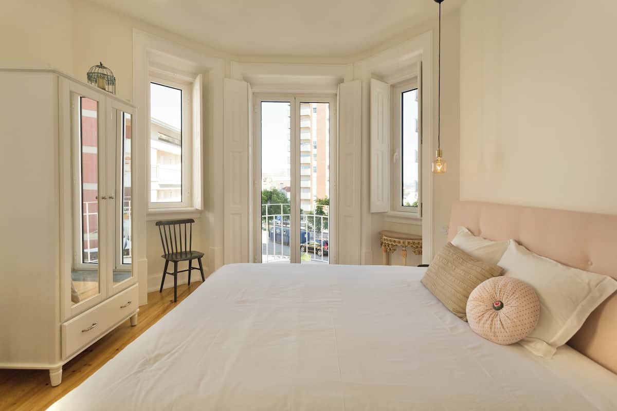 A bedroom with a large bed, a wardrobe, and a chair. There are windows and a balcony visible.