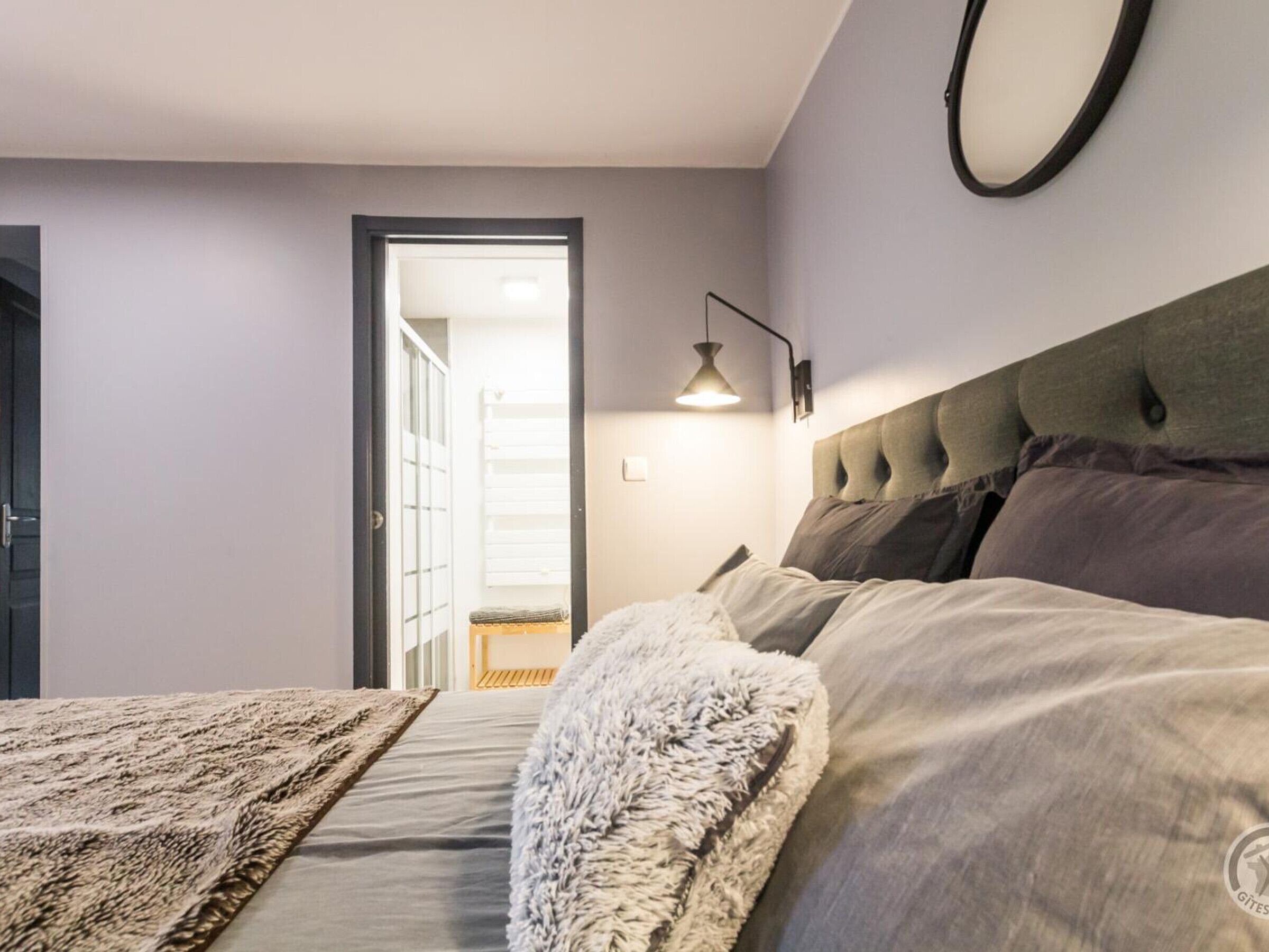 A bedroom with a bed, a nightstand, a wall-mounted lamp, and a door leading to a hallway.