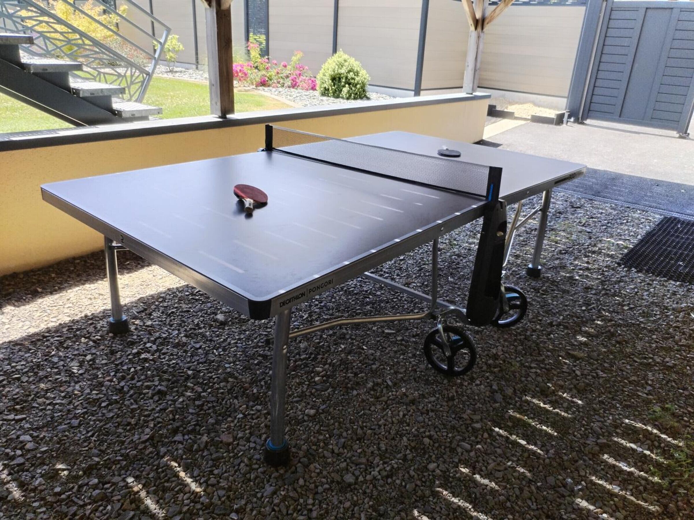 An outdoor ping pong table with a net, set on a gravel surface, with a metal frame and wheels for mobility.