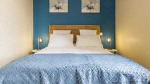 A bedroom with a blue wall, a wooden headboard, two white pillows, a patterned blue bedspread, and two bedside lamps. - Cozy House with Sunny Terrace in Cuves (Cuves)