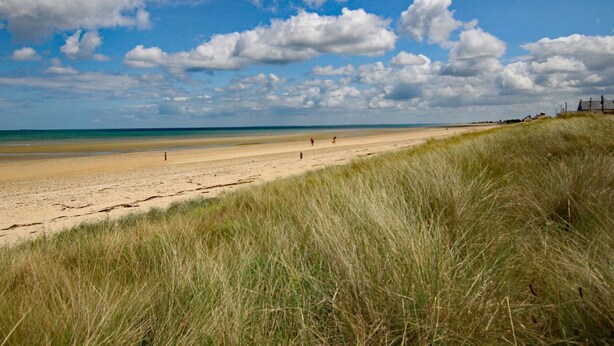 Une plage avec un ciel dégagé et quelques nuages, une rive sablonneuse et de petites dunes herbeuses.