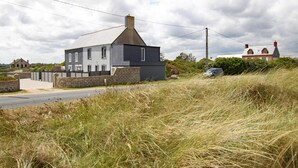 Une maison à deux étages, avec un toit gris et des fondations en pierre, entourée d’herbe haute et sous un ciel nuageux.