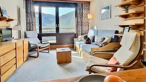 A cozy living room with a sofa, armchairs, a coffee table, and a desk with a lamp. A large window offers a view of mountains.