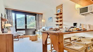 A cozy room with a wooden dining table, chairs, a sofa, a TV mounted on the wall, and a large window with a view of greenery.