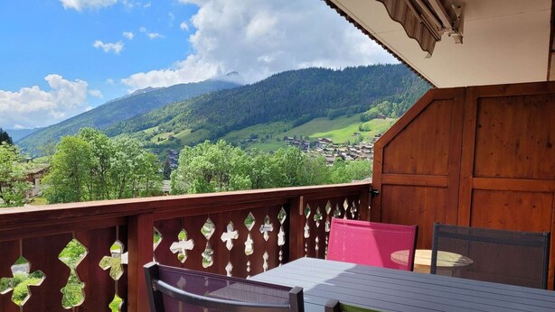 A wooden balcony with a table and chairs, overlooking a valley with houses and mountains. - Beautiful 4-pers. apartment with balcony and garage in Grand-Bornand (Le Grand-Bornand)
