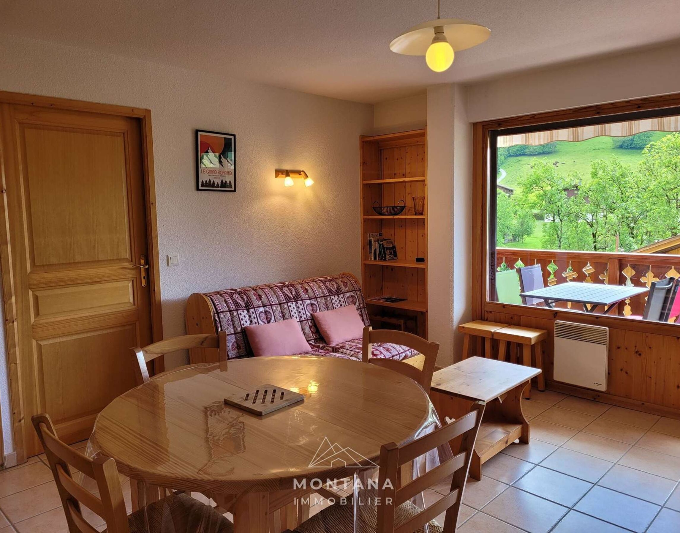 A cozy room with a round wooden table, a sofa, a bookshelf, and a window with a view of greenery.