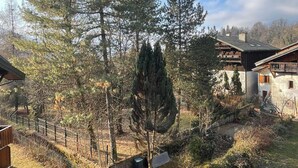 Ett bostadsområde med en stor tall, en träveranda och en byggnad i sten med ett sluttande tak.