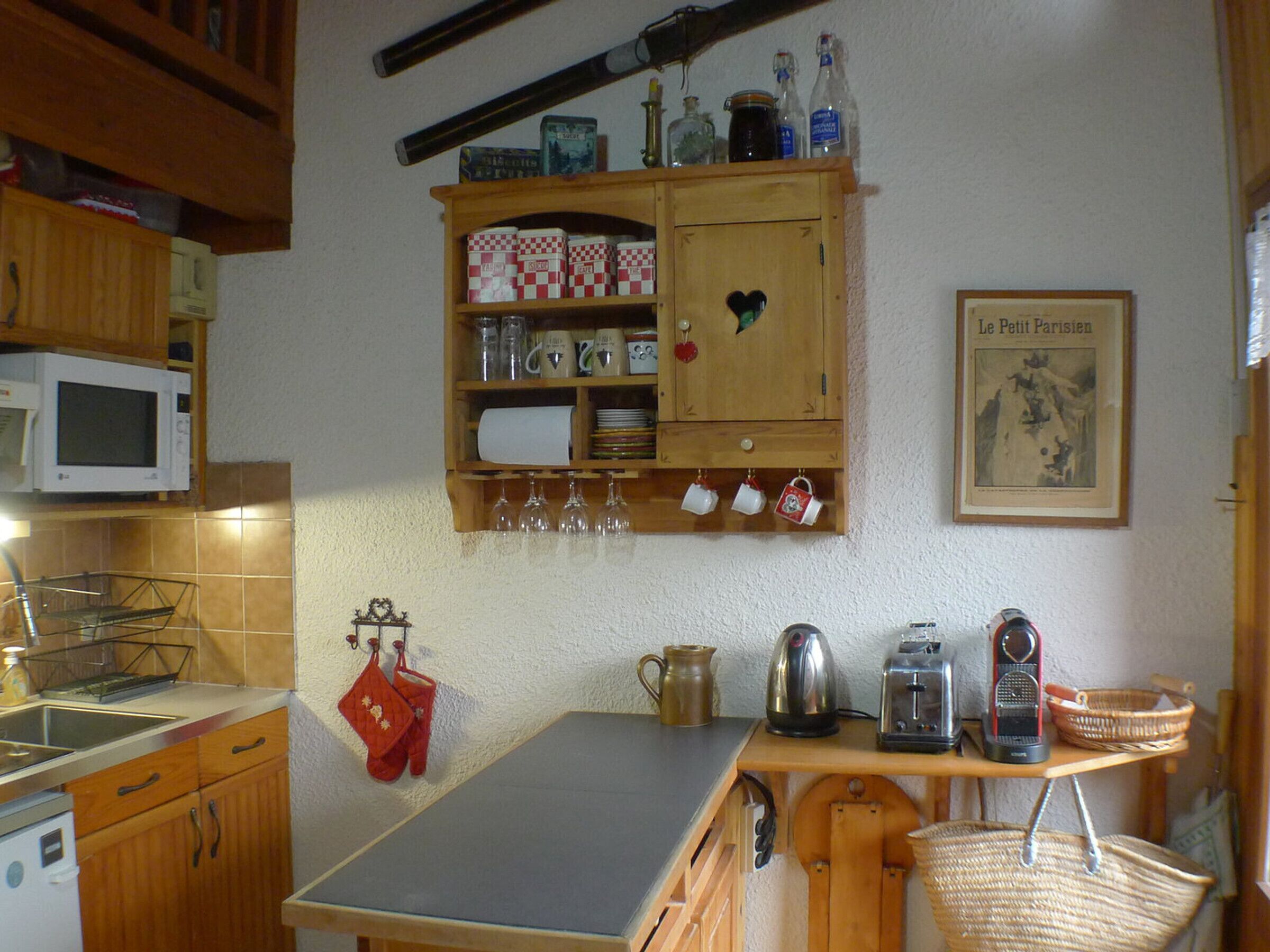 A kitchen with wooden cabinets, a microwave, a kettle, a toaster, and a shelf with jars and glasses.