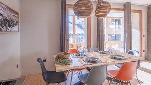 A dining area with a wooden table, black and orange chairs, and a view of a building through large windows. - Charming 3-room apartment with balcony and wifi in Huez (Huez)