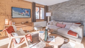 A modern living room with a stone wall, a television displaying a skier, and a cozy seating area with a sofa and armchairs. - Charming 3-room apartment with balcony and wifi in Huez (Huez)