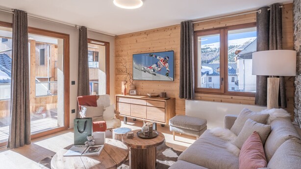 A modern living room with a wooden interior, a large window with a view of the outside, a television mounted on the wall, and a cozy seating area with a sofa and a coffee table. - Charming 3-room apartment with balcony and wifi in Huez (Huez)