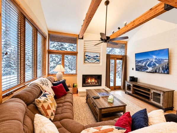 A cozy living room with a fireplace, large windows, a sofa, and a television.