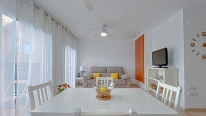 Un coin repas avec une table et des chaises blanches, un ventilateur de plafond, un canapé avec des coussins, un téléviseur posé sur un meuble et une horloge murale.