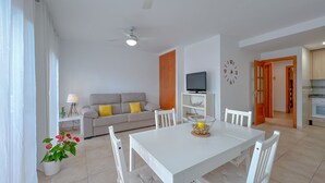 Un salon moderne avec un canapé blanc, une table à manger et des chaises. On y trouve un téléviseur posé sur un meuble, une horloge au mur et un ventilateur de plafond.