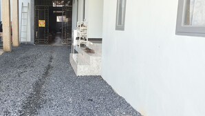A narrow alleyway with a gravel path leading to a gated entrance. A white shelf with shoes is visible on the right. - Hotel O Pondok Tawing Rumbih (Tamban)