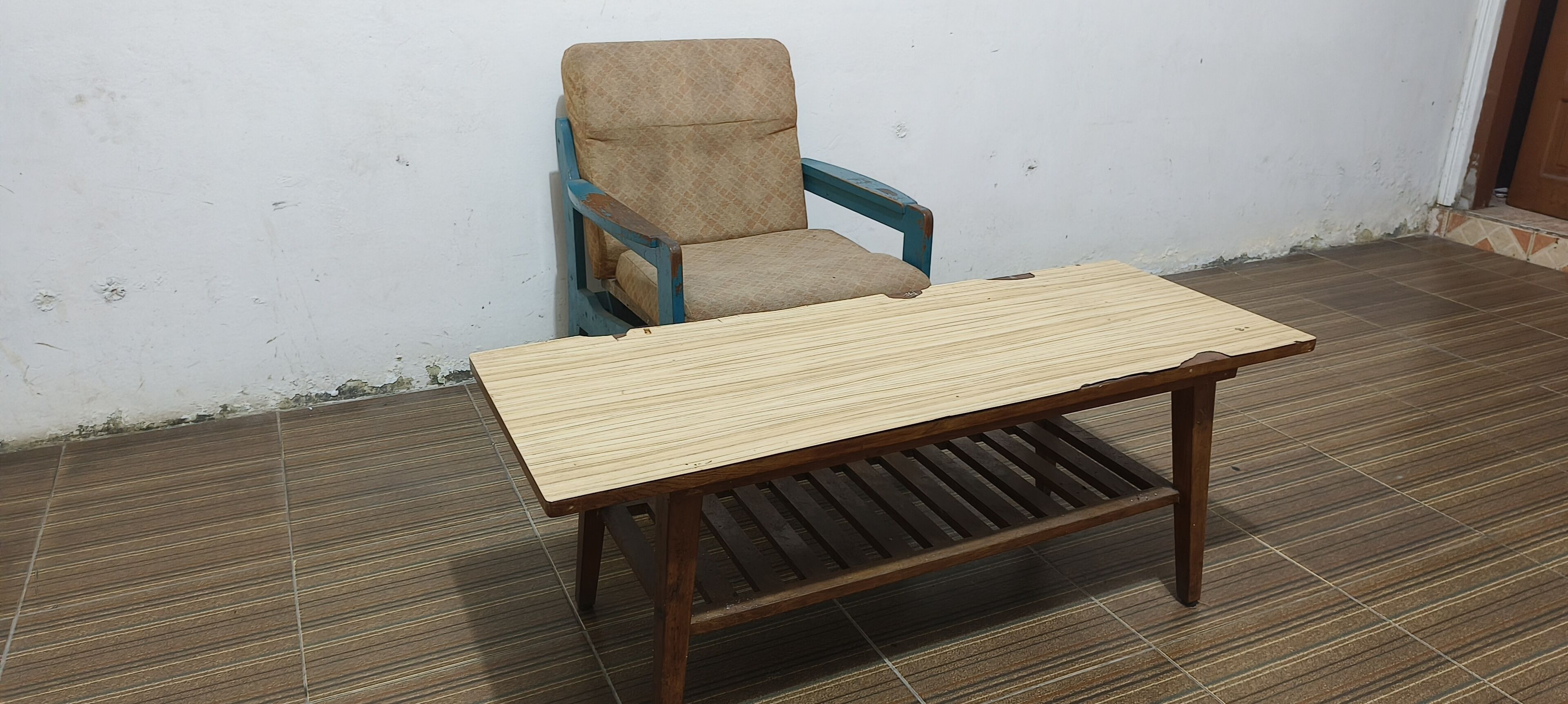 A wooden table with a slatted design, a blue chair with a beige cushion, and a tiled floor.