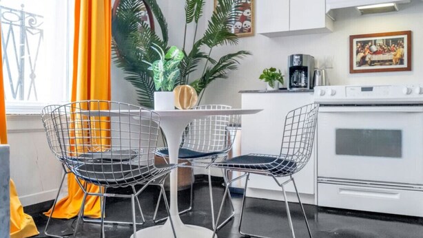 Een moderne keuken met witte kasten, een fornuis en een koffiezetapparaat. Er is een eettafel met draadstoelen en een potplant.