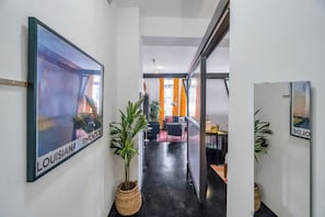 A hallway with a framed picture of a building, a potted plant, and a glimpse of a seating area with orange curtains.