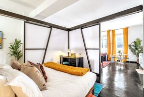 A bedroom with a bed, a nightstand, a lamp, and a view of a dining area with orange curtains.