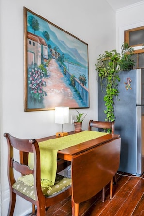 A dining area with a table, chairs, a painting, a lamp, a potted plant, and a refrigerator.