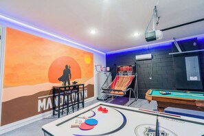 Una habitación con mesa de billar, mesa de ping pong, aro de baloncesto y un mural con un atardecer.