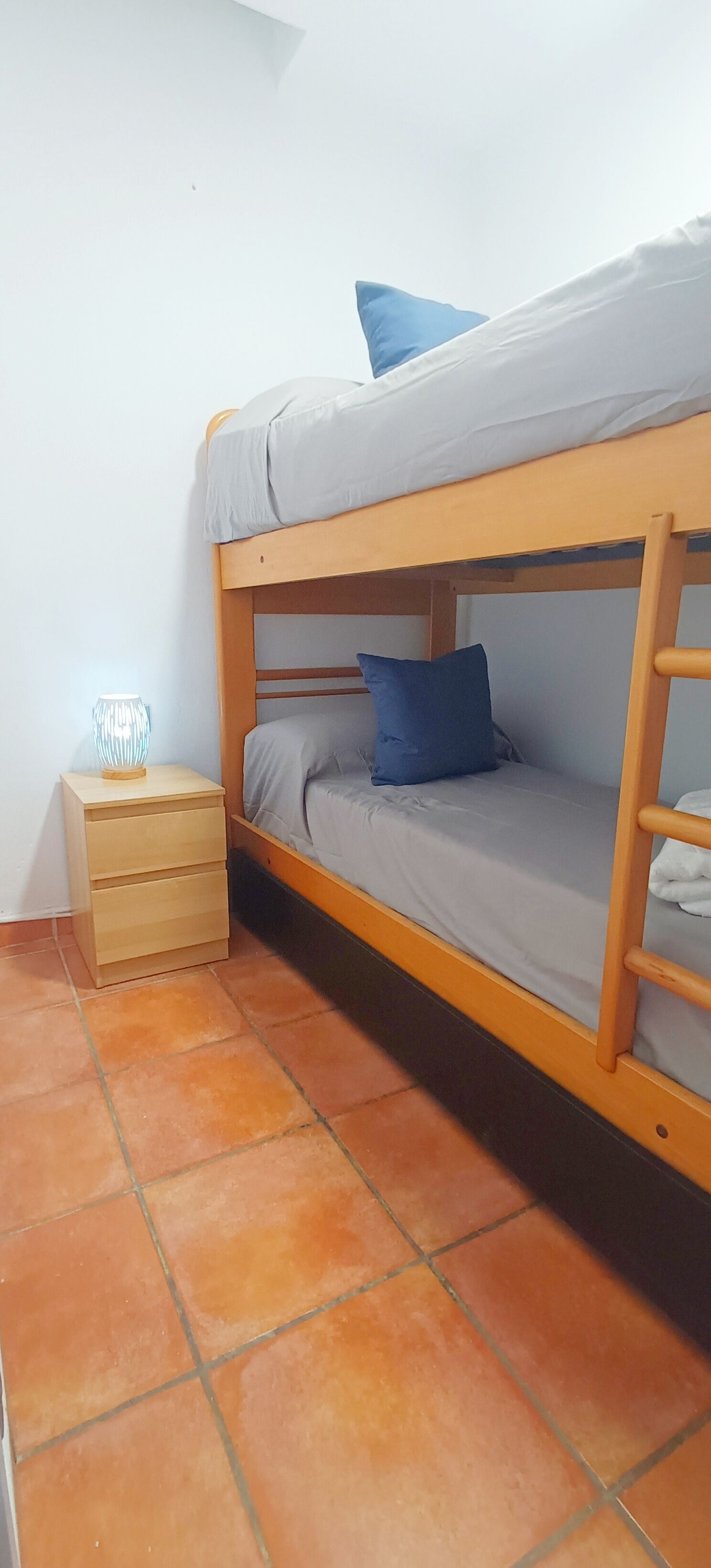 Una cama litera con estructura de madera, una mesita de noche con cajones y una lámpara.