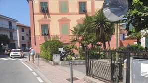 Une vue de rue avec un immeuble dont la façade est rouge et les volets verts, un trottoir bordé d’une grille métallique et une voiture garée.