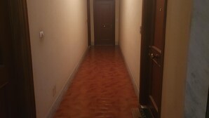 Un couloir étroit, avec des portes en bois de chaque côté, un interrupteur sur le mur de gauche et une porte au fond.