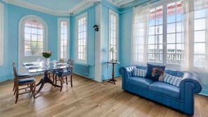 Una habitación con una mesa de comedor y sillas, un sofá azul y ventanas grandes con cortinas blancas.