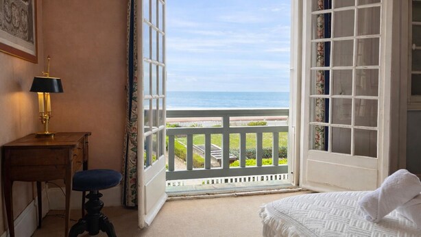 Una habitación con una cama, un escritorio con lámpara y vista al océano por una puerta abierta.