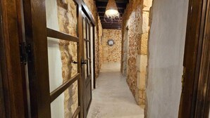 Um corredor estreito com paredes de pedra e portas de madeira, dando acesso a um quarto com um relógio na parede.