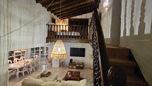 Uma sala de estar ampla com teto alto, uma área de jantar com mobiliário branco e uma escada com corrimão de madeira.