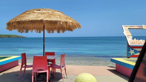 A beachside seating area with a straw umbrella, red chairs, and a small table.