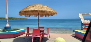 A beachside seating area with a straw umbrella, red chairs, and a small table.