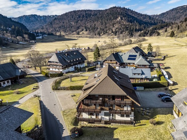 A traditional chalet-style building with a sloped roof, a wooden deck, and a small parking area.