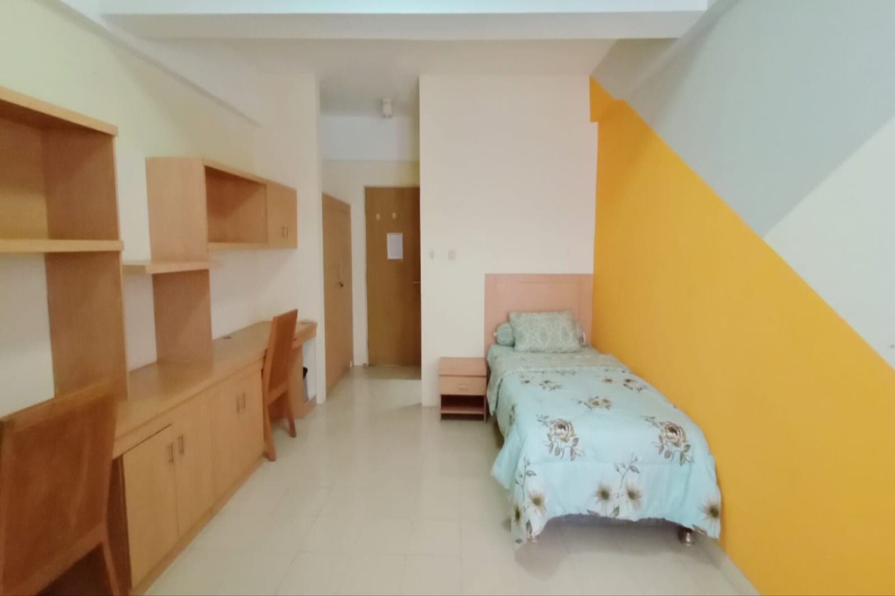 A room with a bed, a desk, and a chair. There is a door leading to another room.