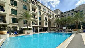 Une piscine entourée de chaises longues et de palmiers, le tout bordé d’un immeuble à plusieurs étages doté de balcons.