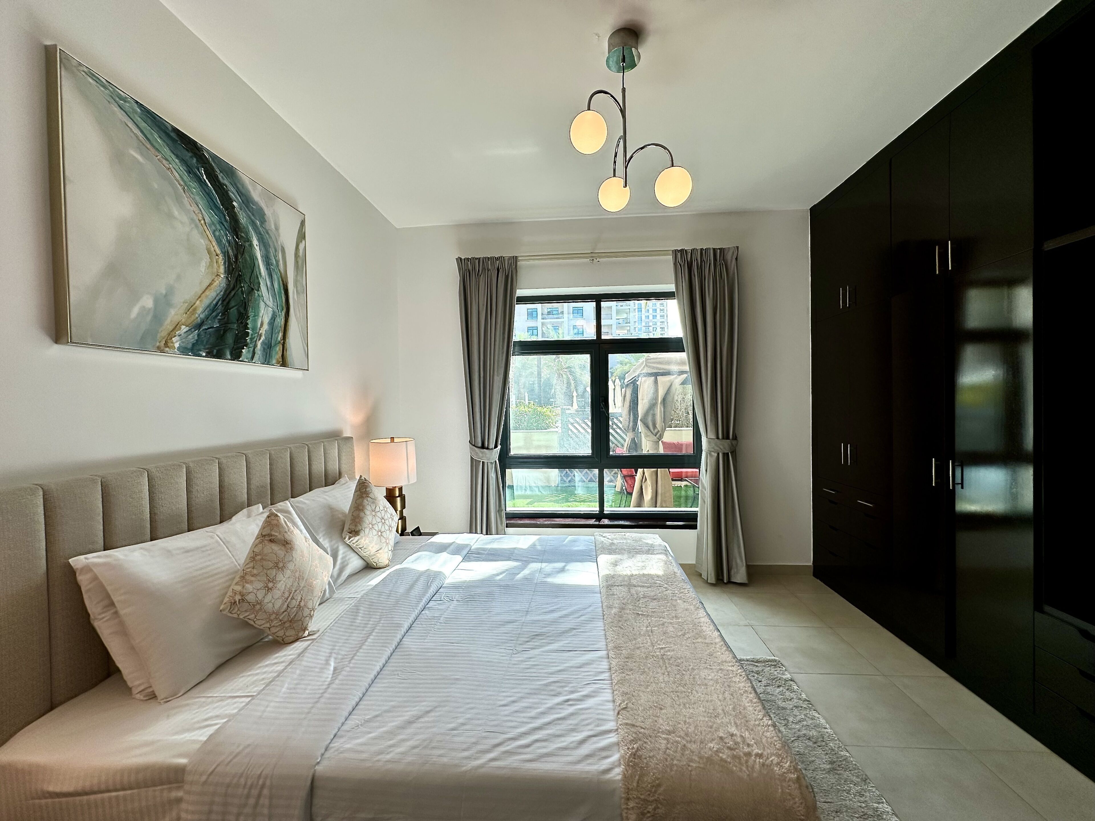 A bedroom with a bed, a bedside lamp, a large abstract painting, a window with curtains, and a view of the outside.